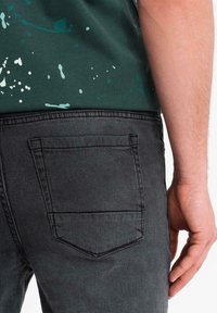 Jeans di denim neri con design della tasca posteriore, abbinati a una maglietta verde scuro con macchie di vernice di colore chiaro e scollo rotondo.