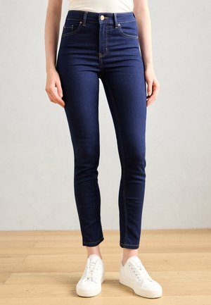 Dunkelblaue, hoch taillierte Jeans im Slim-Fit mit goldenem Nähten, Fronttaschen und einem weißen Knopf. Kombiniert mit weißen Sneakers.