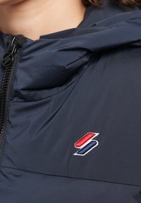 Nærbillede af en marineblå jakke med lynlås og hætte, samt et lille rødt og blåt broderet emblem på brystet nær kravebenet.