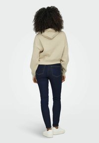 Beige verkürzter Hoodie mit Kordelzugkapuze, kombiniert mit dunkelblauen Skinny Jeans und weißen Sneakers, von hinten betrachtet.