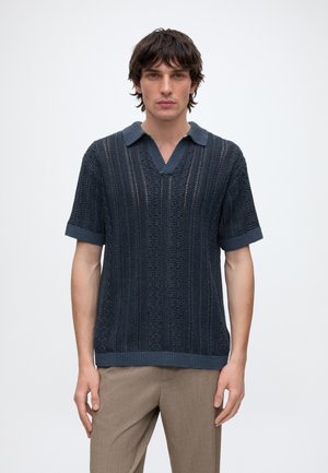 Giovane uomo che indossa una polo in maglia blu navy a maniche corte e pantaloni beige, in piedi davanti a uno sfondo bianco uniforme.