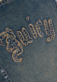 Υφασμα denim με τη λέξη "Juicy" ραμμένη με ξεφτισμένα υφασμάτινα γράμματα διακοσμημένα με μικρές πέρλες.