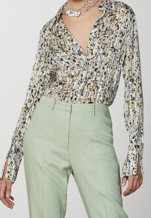 Blouse à manches longues avec un imprimé abstrait aux accents jaunes et noirs, associée à un pantalon taille haute vert clair sur mesure et un collier clair épais.