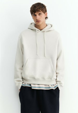 Jeune homme portant un sweat à capuche beige clair avec une poche avant et une chemise rayée en dessous, debout devant un fond blanc uni.