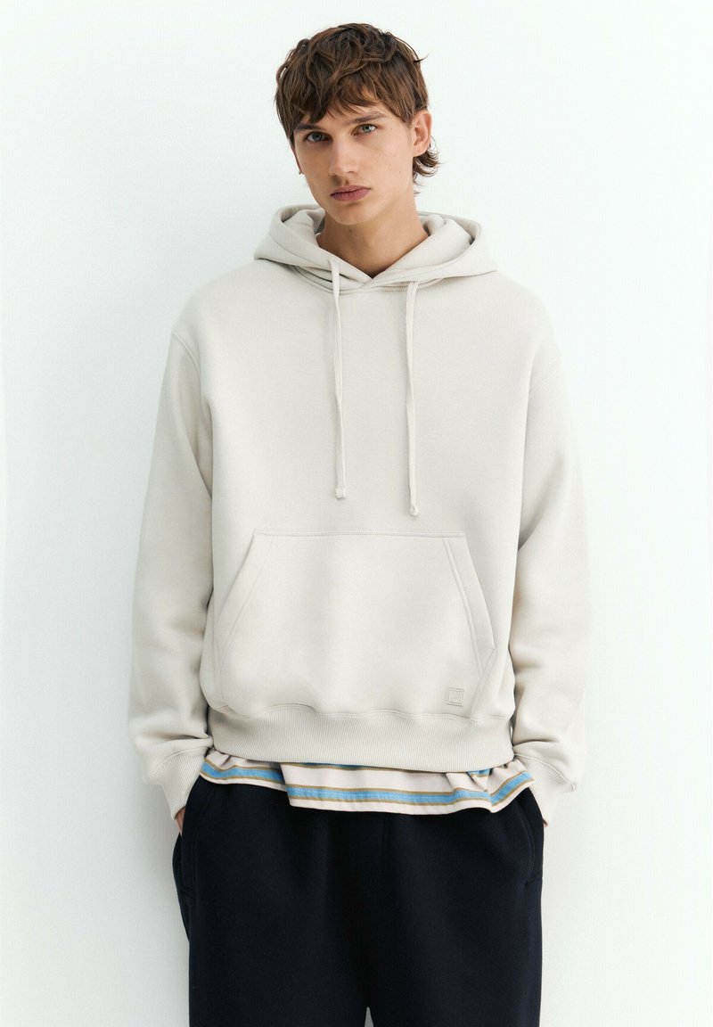 Jeune homme portant un sweat à capuche beige clair avec une poche avant et une chemise rayée en dessous, debout devant un fond blanc uni.