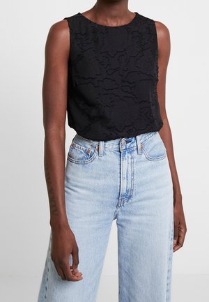 Hihaton musta teksturoitu kukkakuvioinen toppi, jota käyttää korkeavyötäröisten vaaleansinisien farkkujen kanssa tummaihoinen henkilö, joka seisoo.