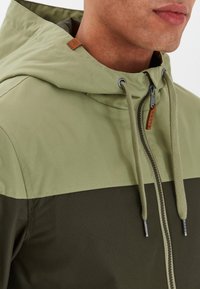 Khaki bunda s kapucňou s dvoma odtieňmi, svetlozelenou hornou časťou a tmavozelenou dolnou časťou, doplnená o sťahovaciu šnúrku a detaily so zipsom.