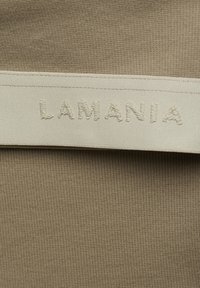 Beeziv värv ja ribaline tekstuur; kontrastne kreemikasvalge silt, millel on sõna "LAMANIA" reljeefse tikandiga õmbluse servas.