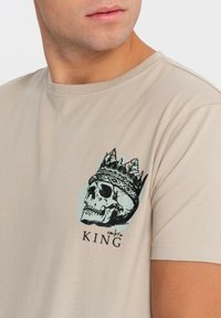 Béžové bavlněné tričko s grafikou černé lebky s korunou, pod designem je text "ombre KING" v černé barvě.
