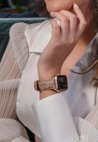 Smartwatch o prostokątnym czarnym wyświetlaczu, złotej ramce i wzorzystym brązowym pasku. Część dłoni opiera się o białą tkaninę mankietu.
