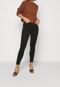 Brun stickad korttröja med ribbad textur, i kombination med svarta tight jeans med texturerade paneler på låren och högklackade sandaler.