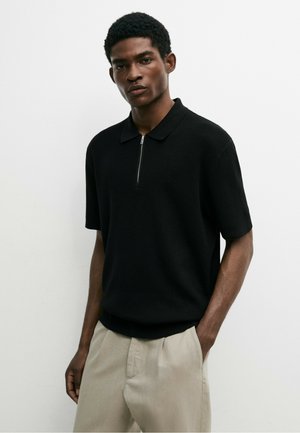 Mann trägt ein schwarzes kurzärmeliges Strick-Poloshirt mit Frontreißverschluss und beigen Hosen, steht vor einem schlichten weißen Hintergrund.