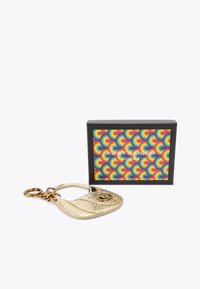 Portachiavi in oro a forma di mini borsa con superficie texture e un elemento decorativo. Scatola colorata con motivi e testo sullo sfondo.