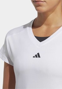 Bílé atletické tričko s krátkým rukávem a výstřihem do V, na hrudi s černým logem Adidas. Hladká struktura látky, minimalistické designové detaily.