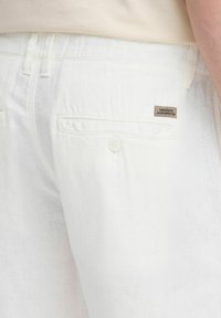 White linen pants with a textured finish, featuring a single back pocket with a button and a small fabric tag sewn on.