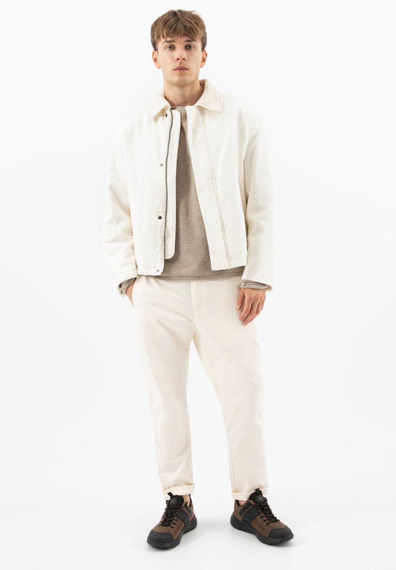 Jeune homme portant une veste beige clair, un pull beige, un pantalon crème et des baskets marron, debout les mains dans les poches sur un fond blanc.