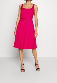 Fuchsiové šaty bez rukávů se čtvercovým výstřihem, těsným korzetem a rozšířenou sukní; obsahují tenký odpovídající pásek a hladkou texturu látky.