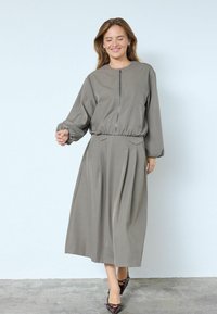 Pelēkas krāsas kleita ar gariem piedurknēm, priekšpusē ar rāvējslēdzēju, elastīgu vidukli un plisētu svārku daļu. Gluda auduma, brīva piegriezuma, ar apaļu kakla izgriezumu un minimālu kabatu skaitu.