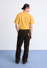 Gul bomulds t-shirt, korte ærmer, parret med mørkebrun corduroy bukser med to baglommen og en logo patch. Brune sko.