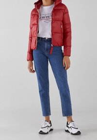 Rød pufferjakke med hætte og lynlås, parret med lyseblå straight-leg jeans. Sorte og hvide sporty sneakers fuldender outfittet.