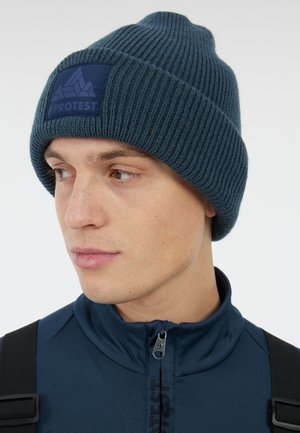 Dark blue ribbed knit beanie with a matching logo patch. Worn with a high-collar jacket, highlighting the hat's texture and fit.