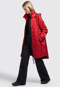 Chaqueta impermeable roja con capucha, bolsillos frontales y cintura ceñida. Combinada con pantalones anchos negros y calzado negro.