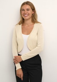 Vaalea beige lyhyt keskustelupaita, jossa on pitkät hihat, pehmeä rakenne ja sileä pinta, puettuna valkoisen paidan ja mustien housujen päälle.