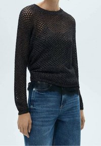 Zwarte gebreide trui met een open geweven ontwerp, lange mouwen en een geribbelde halslijn. Gecombineerd met blauwe spijkerbroeken.