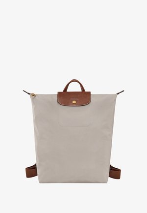 Beige nylon backpack with a textured brown leather flap and handle, gold hardware, and adjustable brown straps. Minimalist design.
