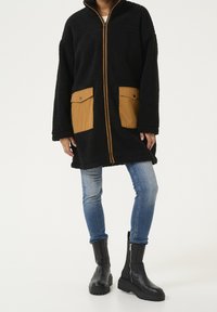 Czarny płaszcz z polaru z brązowymi kieszeniami naszywanymi, zamkiem z przodu i wysokim kołnierzem. W połączeniu z niebieskimi dżinsami i czarnymi botkami za kostkę.
