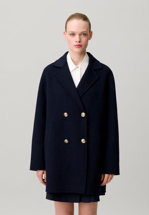 Donna che indossa un cappotto doppiopetto blu navy scuro con bottoni dorati sopra una camicia bianca con colletto e una gonna scura, su uno sfondo chiaro e uniforme.