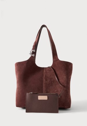 Braune Wildleder-Shopper-Tasche mit silberner Schnalle und passendem, strukturiertem Leder-Reißverschluss-Portemonnaie vorne auf weißem Hintergrund befestigt.