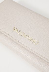 Portafoglio in pelle grigia chiara con superficie texturizzata e logo dorato "VALENTINO". Presenta un design a pieghe e cuciture visibili in evidenza.