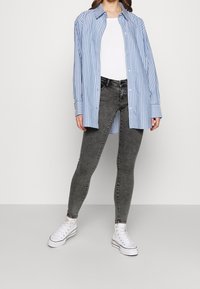 Camisa de botones de rayas azul y blanca con mangas largas, combinada con jeans ajustados grises y deportivas blancas, de pie contra un fondo liso.