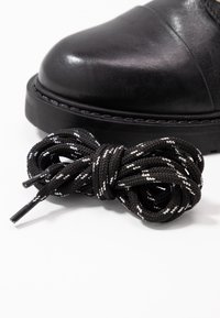 Bota de cuero negra con punta redondeada, acompañada de cordones de patrón negro y blanco enrollados cercanos. Textura suave en la superficie de la bota.