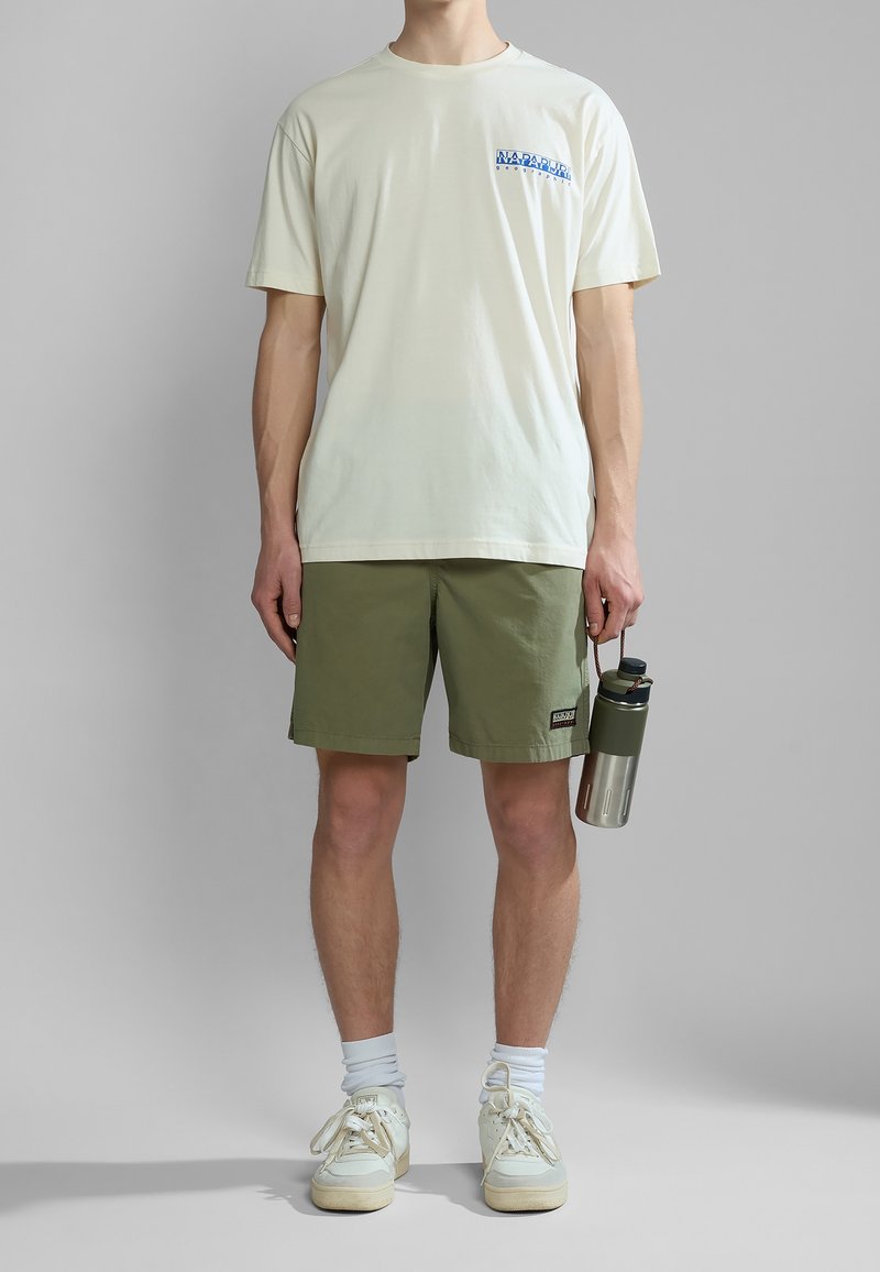 Homme portant un t-shirt blanc Napapijri, un short vert olive, des chaussettes blanches, des baskets, tenant une bouteille d'eau verte en acier inoxydable.