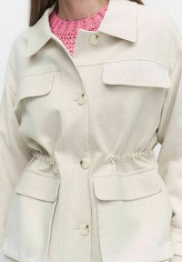 Ljusbeige jacka i tyg med en texturerad yta. Har dragsko i midjan, stora framfickor och knappstängning.