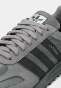 Αθλητικό παπούτσι από ανοιχτό γκρι σουέντ και δίχτυ με σκούρες γκρι ρίγες, υφασμάτινες λεπτομέρειες και ασημένια ετικέτα με το λογότυπο στη γλώσσα. Τα κορδόνια είναι παστέλ γκρι.