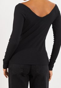 Schwarzes, geripptes Langarmtop mit weit ausgeschnittenem V-Ausschnitt hinten, aus elastischem Stoff, kombiniert mit dunklen Jeans.