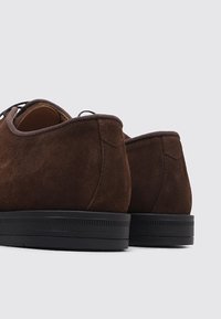Par de zapatos de ante marrón de cordones con suelas de goma negra, vistos desde detrás sobre un fondo claro y uniforme.