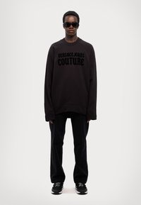 Crni oversized dukserica s logotipom "VERSACE JEANS COUTURE", uparen s crnim hlačama i crnim tenisicama, postavljenim nasuprot sivoj pozadini.