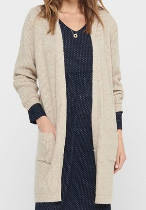 Femme portant un cardigan beige en maille par-dessus une robe bleu marine à pois avec des manches longues, les mains dans les poches du cardigan, et un collier pendentif.