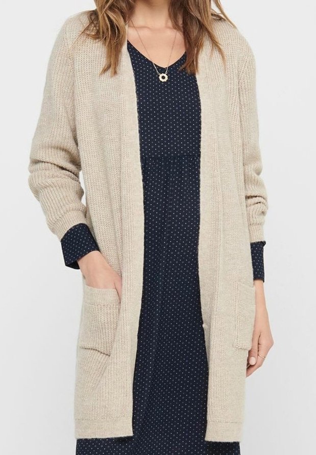 Femme portant un cardigan beige en maille par-dessus une robe bleu marine à pois avec des manches longues, les mains dans les poches du cardigan, et un collier pendentif.
