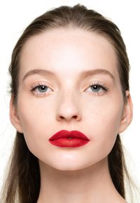 Primer plano de una cara con piel suave, mejillas pecosas, labios rojos mate intensos y un maquillaje natural que enfatiza los ojos claros y las largas pestañas.