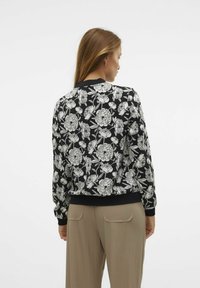 Černá bomber bunda s bílým květinovým vzorem, s ribbed manžetami a spodním lemem, v uvolněném střihu. Zobrazený zadní pohled.