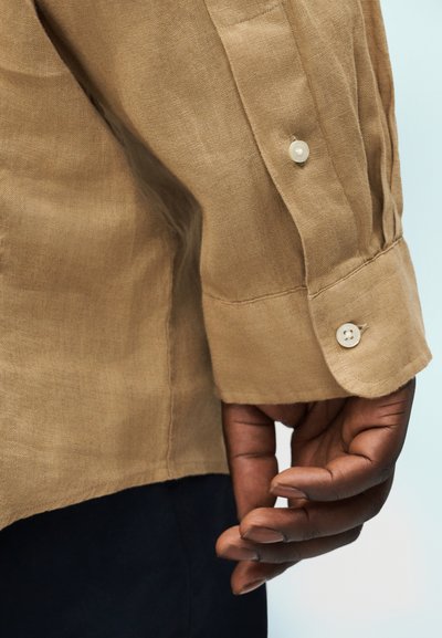 Tamsios odos ranka, atsipalaidavusi prie šono, dėvinti smėlio spalvos ilgomis rankovėmis marškinius su užsegamu rankogaliu ir juodas kelnes, priešais šviesų foną.