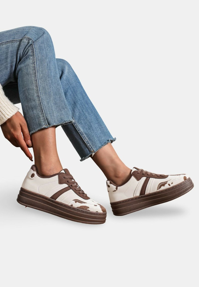 Baskets plateforme blanches et brunes avec une surface texturée, présentant des rayures brunes, des lacets et un motif de tache de vache sur le bout.