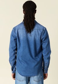 Koszula dżinsowa w średnim niebieskim kolorze z długimi rękawami, mankietami na guziki i zaokrąglonym dolnym brzegiem. Gładka tekstura i minimalne detale przeszyć.