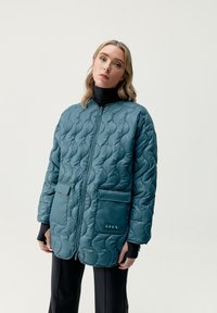 Giacca puffer trapuntata color teal con zip frontale, due tasche a toppa e un accento con logo. Presenta un colletto alto e una vestibilità rilassata.