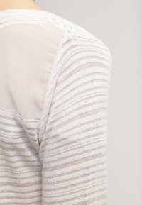 Lehká pruhovaná halenka s jemným, průsvitným zadním panelem. Tkanina má jemnou texturu s vodorovnými liniemi v odstínech béžové a krémové.